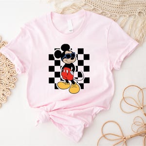 May include: Pink t-shirt with a black and white checkered background and a cartoon image of Mickey Mouse wearing sunglasses. Mickey Mouse is wearing red shorts and black shoes.