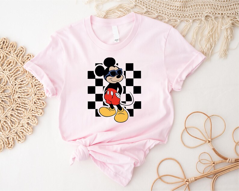 May include: Pink t-shirt with a black and white checkered background and a cartoon image of Mickey Mouse wearing sunglasses. Mickey Mouse is wearing red shorts and black shoes.