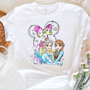 May include: White t-shirt with a colorful illustration of two princesses, Elsa and Anna, from the movie Frozen. The princesses are standing in front of a castle with a Mickey Mouse ear bow above them.