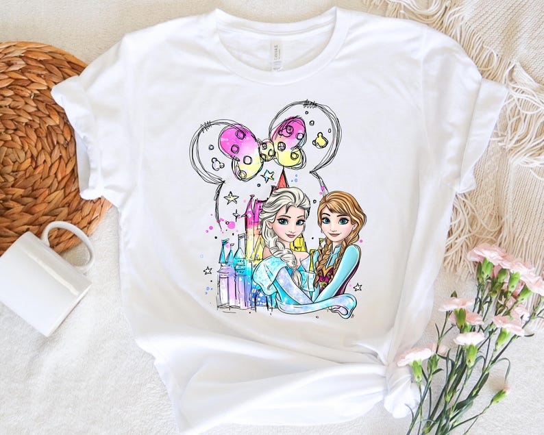 May include: White t-shirt with a colorful illustration of two princesses, Elsa and Anna, from the movie Frozen. The princesses are standing in front of a castle with a Mickey Mouse ear bow above them.