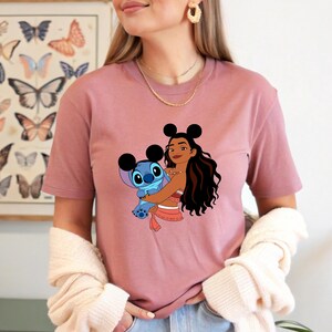 May include: Dusty rose t-shirt featuring a graphic of a cartoon character hugging Stitch. The character has black mouse ears and long dark hair. Stitch is blue. The shirt is worn by a person.