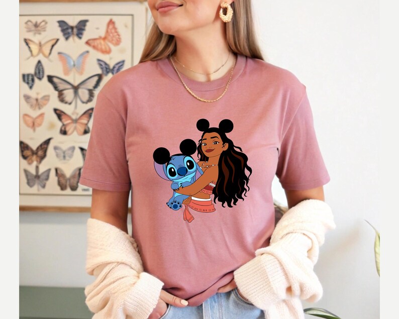 May include: Dusty rose t-shirt featuring a graphic of a cartoon character hugging Stitch. The character has black mouse ears and long dark hair. Stitch is blue. The shirt is worn by a person.