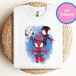May include: White sweatshirt with a vibrant cartoon illustration of Spider-Man characters. The design features Spider-Gwen, Miles Morales, and Spider-Man, set against a blue backdrop. A pink circle with the text "DTF TRANSFER" is in the upper right.