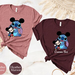 May include: Two t-shirts with a cartoon image of Stitch from Lilo & Stitch holding a small Mickey Mouse. The shirt on the left is heather mauve and the shirt on the right is heather maroon. The maroon shirt has the text "Custom Text" printed below the image.