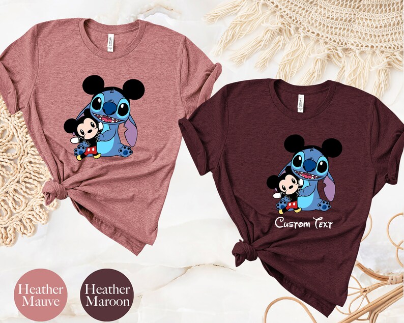 May include: Two t-shirts with a cartoon image of Stitch from Lilo & Stitch holding a small Mickey Mouse. The shirt on the left is heather mauve and the shirt on the right is heather maroon. The maroon shirt has the text "Custom Text" printed below the image.