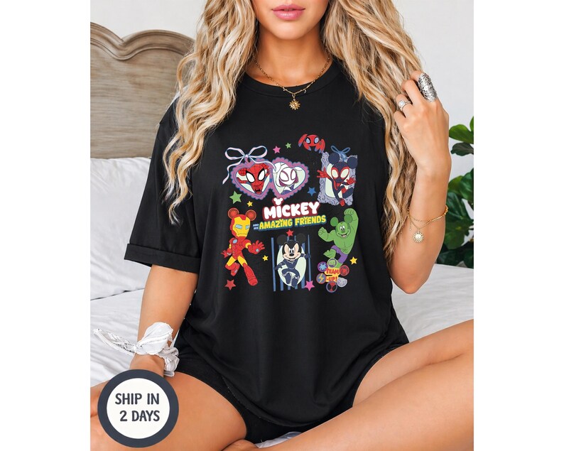 May include: Black t-shirt featuring a colorful graphic of various cartoon characters, including Spider-Man, Mickey Mouse, and Iron Man. The text "Mickey Amazing Friends" is also displayed. The shirt is short-sleeved and appears to be made of a soft material.