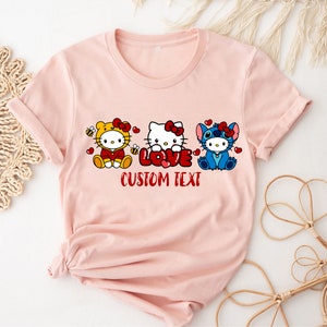 May include: A light pink t-shirt featuring three cartoon characters: a yellow bee-themed character, Hello Kitty, and Stitch. The word "LOVE" is in red, with "CUSTOM TEXT" below. The design is centered on the shirt.