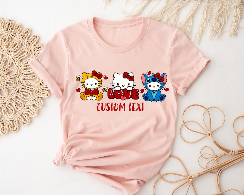 May include: A light pink t-shirt featuring three cartoon characters: a yellow bee-themed character, Hello Kitty, and Stitch. The word "LOVE" is in red, with "CUSTOM TEXT" below. The design is centered on the shirt.