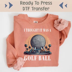 May include: A coral colored sweatshirt with the text "I thought it was a golf ball" printed on the front. The graphic features a cartoon illustration of the Spaceship Earth attraction at Epcot, a theme park in Orlando, Florida.