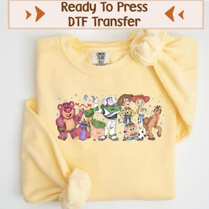 Peut inclure: Un sweat-shirt jaune clair avec un motif coloré de personnages de dessin animé. Le motif comprend un ours, un dinosaure, un ranger de l'espace, une cow-girl, un cow-boy, une tête de pomme de terre, un cheval et un cochon. Le texte "Ready To Press DTF Transfer" est en haut.