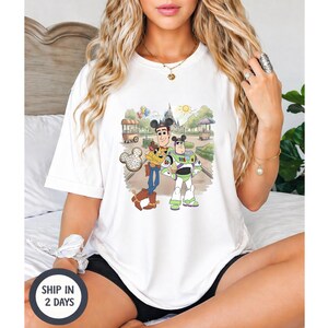 May include: A white short-sleeved t-shirt featuring a graphic print of Woody and Buzz Lightyear wearing Mickey Mouse ears. The characters are standing in a park setting with a castle in the background, accompanied by a Mickey-shaped pretzel icon.