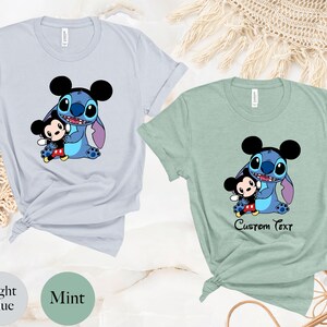 May include: Two t-shirts with a blue Stitch character holding a Mickey Mouse character. The shirt on the left is light blue and the shirt on the right is mint green. The shirt on the right has the text "Custom Text" printed on it.