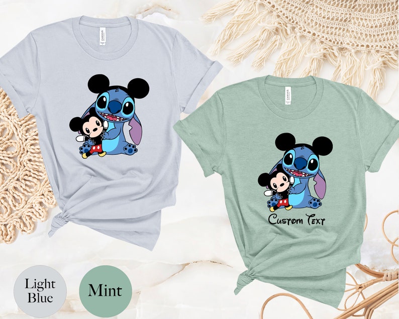 May include: Two t-shirts with a blue Stitch character holding a Mickey Mouse character. The shirt on the left is light blue and the shirt on the right is mint green. The shirt on the right has the text "Custom Text" printed on it.
