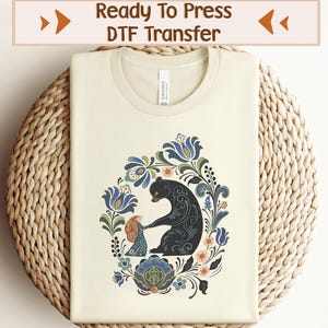 以下が含まれることがあります： 丸いネックラインのクリーム色のTシャツ。黒いクマと人物のフローラルデザインが特徴です。デザインには青、緑、桃色の花が含まれています。「Ready To Press DTF Transfer」というテキストが上部にあります。