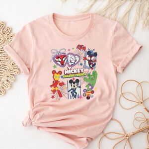May include: A light pink t-shirt featuring a colorful graphic of various Disney characters in superhero costumes. The graphic includes the text "Mickey Amazing Friends". The shirt has short sleeves and a crew neck.