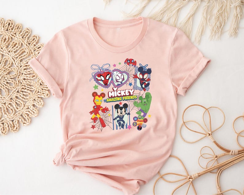 May include: A light pink t-shirt featuring a colorful graphic of various Disney characters in superhero costumes. The graphic includes the text "Mickey Amazing Friends". The shirt has short sleeves and a crew neck.