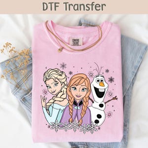 Może przedstawiać: Jasnoróżowa bluza Comfort Colors z transferem DTF Elsy, Anny i Olafa z Krainy Lodu. Grafika zawiera płatki śniegu i tekst "DTF Transfer" na górze. Złoty naszyjnik jest zarzucony na koszulę.
