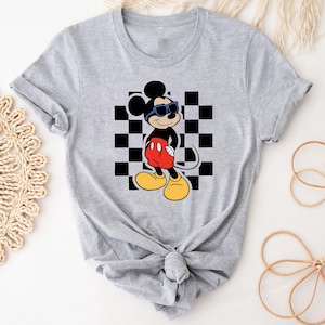 May include: A gray t-shirt with a black and white checkered pattern and a cartoon image of Mickey Mouse wearing sunglasses. Mickey Mouse is wearing red shorts and black shoes.