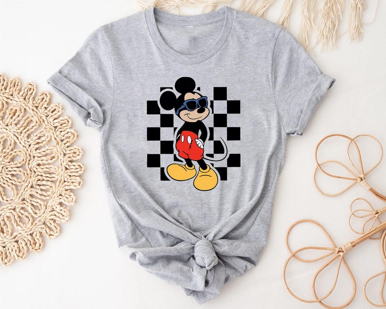 May include: A gray t-shirt with a black and white checkered pattern and a cartoon image of Mickey Mouse wearing sunglasses. Mickey Mouse is wearing red shorts and black shoes.