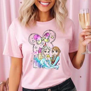 May include: Pink t-shirt with a graphic of two cartoon princesses, Elsa and Anna, standing in front of a castle with a Mickey Mouse ear balloon. The princesses are wearing their signature blue and purple dresses.