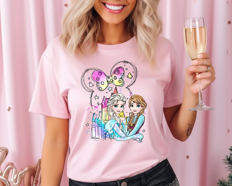 May include: Pink t-shirt with a graphic of two cartoon princesses, Elsa and Anna, standing in front of a castle with a Mickey Mouse ear balloon. The princesses are wearing their signature blue and purple dresses.