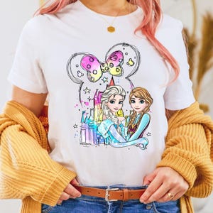 May include: White t-shirt with a colorful illustration of two princesses, Elsa and Anna, standing in front of a castle. The princesses are wearing their signature blue and purple dresses. The illustration is surrounded by a pink bow and stars.