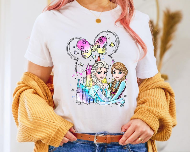 May include: White t-shirt with a colorful illustration of two princesses, Elsa and Anna, standing in front of a castle. The princesses are wearing their signature blue and purple dresses. The illustration is surrounded by a pink bow and stars.