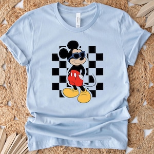 May include: Light blue t-shirt with a black and white checkered pattern and a cartoon image of Mickey Mouse wearing sunglasses and red shorts.