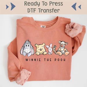 May include: A ready-to-press DTF transfer featuring Winnie the Pooh and his friends. The transfer shows Eeyore, Winnie the Pooh, Piglet, and Tigger. The transfer is on a peach-coloured sweatshirt.