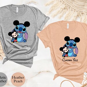 May include: Two t-shirts featuring a blue Stitch character from Lilo & Stitch holding a Mickey Mouse plush. The shirt on the left is heather gray and the shirt on the right is heather peach. The shirt on the right has the text "Custom Text" printed below the image.