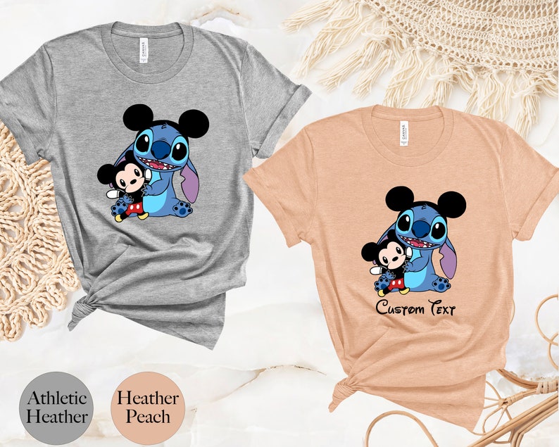 May include: Two t-shirts featuring a blue Stitch character from Lilo & Stitch holding a Mickey Mouse plush. The shirt on the left is heather gray and the shirt on the right is heather peach. The shirt on the right has the text "Custom Text" printed below the image.