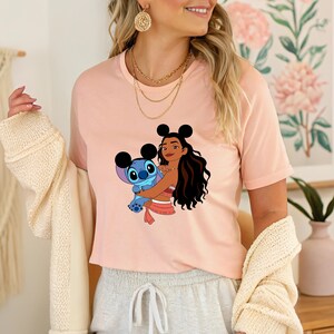 May include: A light pink t-shirt featuring a graphic of a cartoon character embracing another cartoon character. The graphic is in shades of blue, brown, and pink. The shirt is short-sleeved and has a crew neck.
