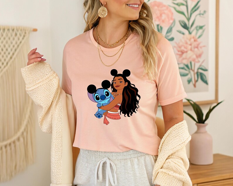 May include: A light pink t-shirt featuring a graphic of a cartoon character embracing another cartoon character. The graphic is in shades of blue, brown, and pink. The shirt is short-sleeved and has a crew neck.