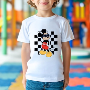 May include: White t-shirt with a black and white checkered background and a cartoon image of Mickey Mouse wearing sunglasses. Mickey Mouse is wearing red shorts and black shoes.