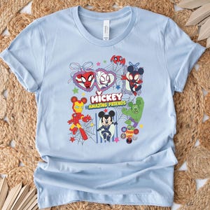 May include: Light blue t-shirt featuring a colorful graphic of Mickey Mouse and other Marvel characters. The design includes Spider-Man, Iron Man, and the Hulk, with the text "Mickey Amazing Friends". The shirt is made of soft material.