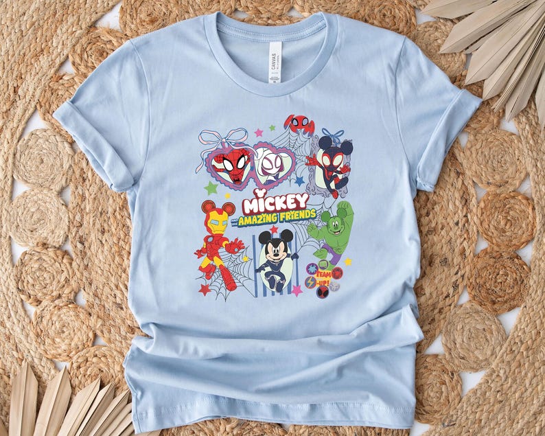 May include: Light blue t-shirt featuring a colorful graphic of Mickey Mouse and other Marvel characters. The design includes Spider-Man, Iron Man, and the Hulk, with the text "Mickey Amazing Friends". The shirt is made of soft material.