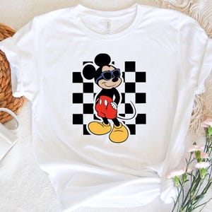 May include: White t-shirt with a black and white checkered background and a cartoon image of Mickey Mouse wearing sunglasses. Mickey Mouse is wearing red shorts and yellow shoes.