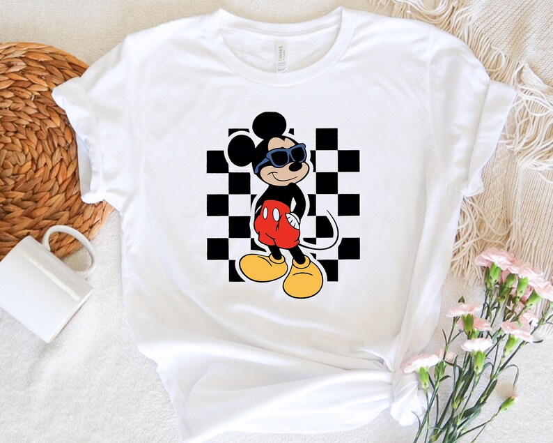 May include: White t-shirt with a black and white checkered background and a cartoon image of Mickey Mouse wearing sunglasses. Mickey Mouse is wearing red shorts and yellow shoes.