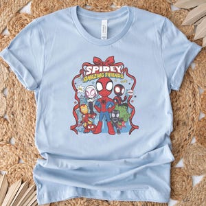 以下が含まれることがあります： スパイダーマンと他のマーベルキャラクターのグラフィックが特徴のライトブルーのTシャツ。デザインには、赤いリボンのようなフレームに「Spidey Amazing Friends」というテキストが含まれています。シャツは柔らかい素材で作られています。