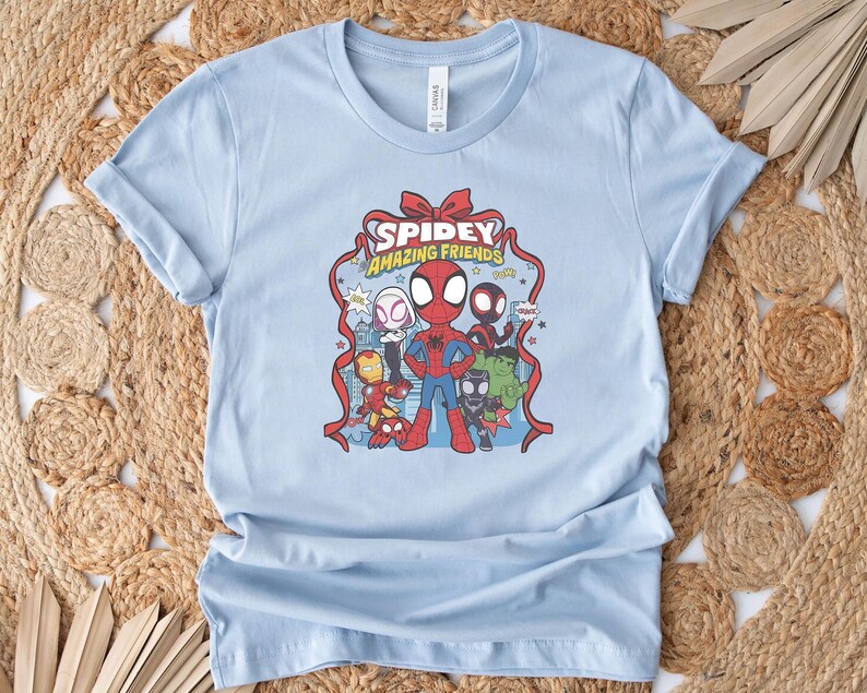以下が含まれることがあります： スパイダーマンと他のマーベルキャラクターのグラフィックが特徴のライトブルーのTシャツ。デザインには、赤いリボンのようなフレームに「Spidey Amazing Friends」というテキストが含まれています。シャツは柔らかい素材で作られています。