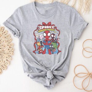 以下が含まれることがあります： スパイダーマンと仲間たちのカラフルなグラフィックがプリントされたグレーのTシャツ。「Spidey Amazing Friends」の文字が赤と黄色でデザインされています。スパイダーマン、アイアンマン、その他のキャラクターが登場する漫画です。