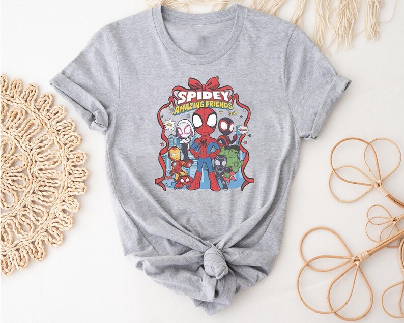 以下が含まれることがあります： スパイダーマンと仲間たちのカラフルなグラフィックがプリントされたグレーのTシャツ。「Spidey Amazing Friends」の文字が赤と黄色でデザインされています。スパイダーマン、アイアンマン、その他のキャラクターが登場する漫画です。
