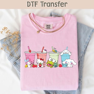 May include: A light pink t-shirt featuring a colorful graphic of cartoon characters with bubble tea drinks. The text "DTF Transfer" is at the top. The shirt is paired with blue jeans and a necklace.
