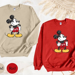 May include: Two sweatshirts with a cartoon drawing of Mickey Mouse. One sweatshirt is sand colored and the other is red.