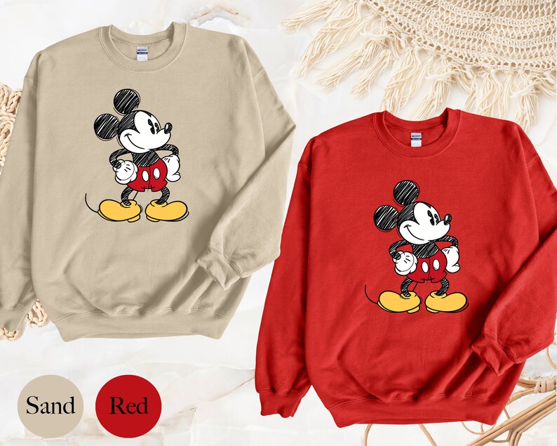 May include: Two sweatshirts with a cartoon drawing of Mickey Mouse. One sweatshirt is sand colored and the other is red.