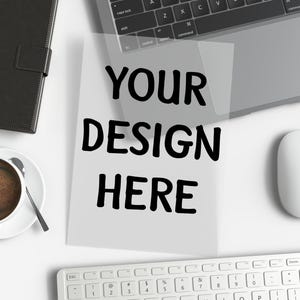 Peut inclure: Un bureau blanc avec un carnet en cuir noir, un ordinateur portable, une tasse à café blanche avec du café, une souris sans fil blanche, un clavier blanc et une feuille de papier avec le texte "YOUR DESIGN HERE" imprimé dessus.