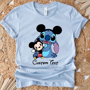 May include: Light blue t-shirt with a cartoon image of Stitch and Mickey Mouse. The text "Custom Text" is printed below the image.