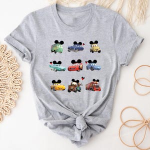 Puede incluir: Una camiseta gris con varios personajes de Cars que llevan orejas de Mickey Mouse. Los personajes incluyen a Mater, Rayo McQueen y algunos otros.