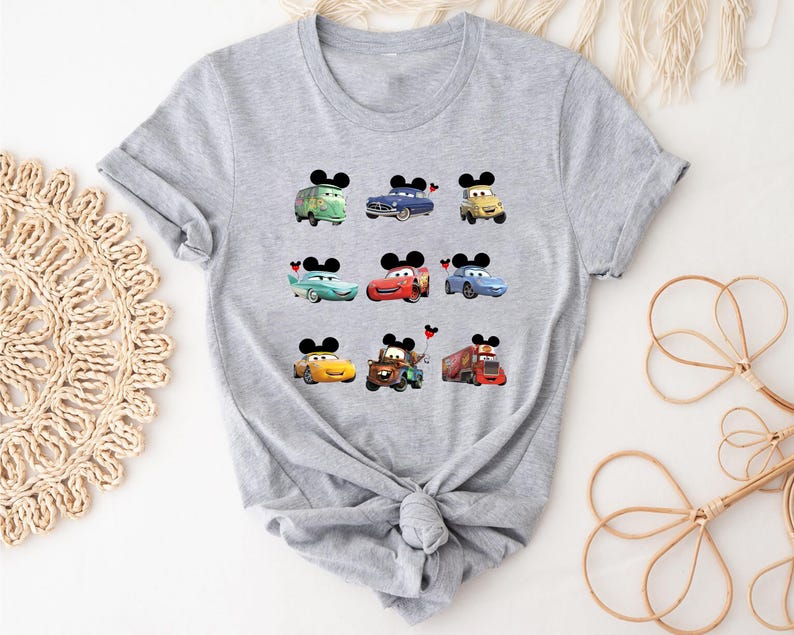 Puede incluir: Una camiseta gris con varios personajes de Cars que llevan orejas de Mickey Mouse. Los personajes incluyen a Mater, Rayo McQueen y algunos otros.