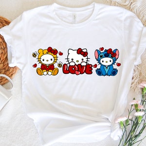 May include: White t-shirt featuring three cartoon characters: Hello Kitty in a tiger costume, Hello Kitty holding the word "LOVE", and Stitch. Each character has a red bow and is surrounded by hearts.